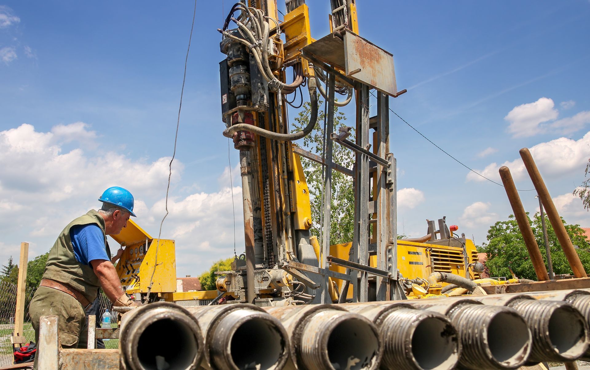 Workers operate a drilling rig beside stacked concrete pipes under a blue sky.