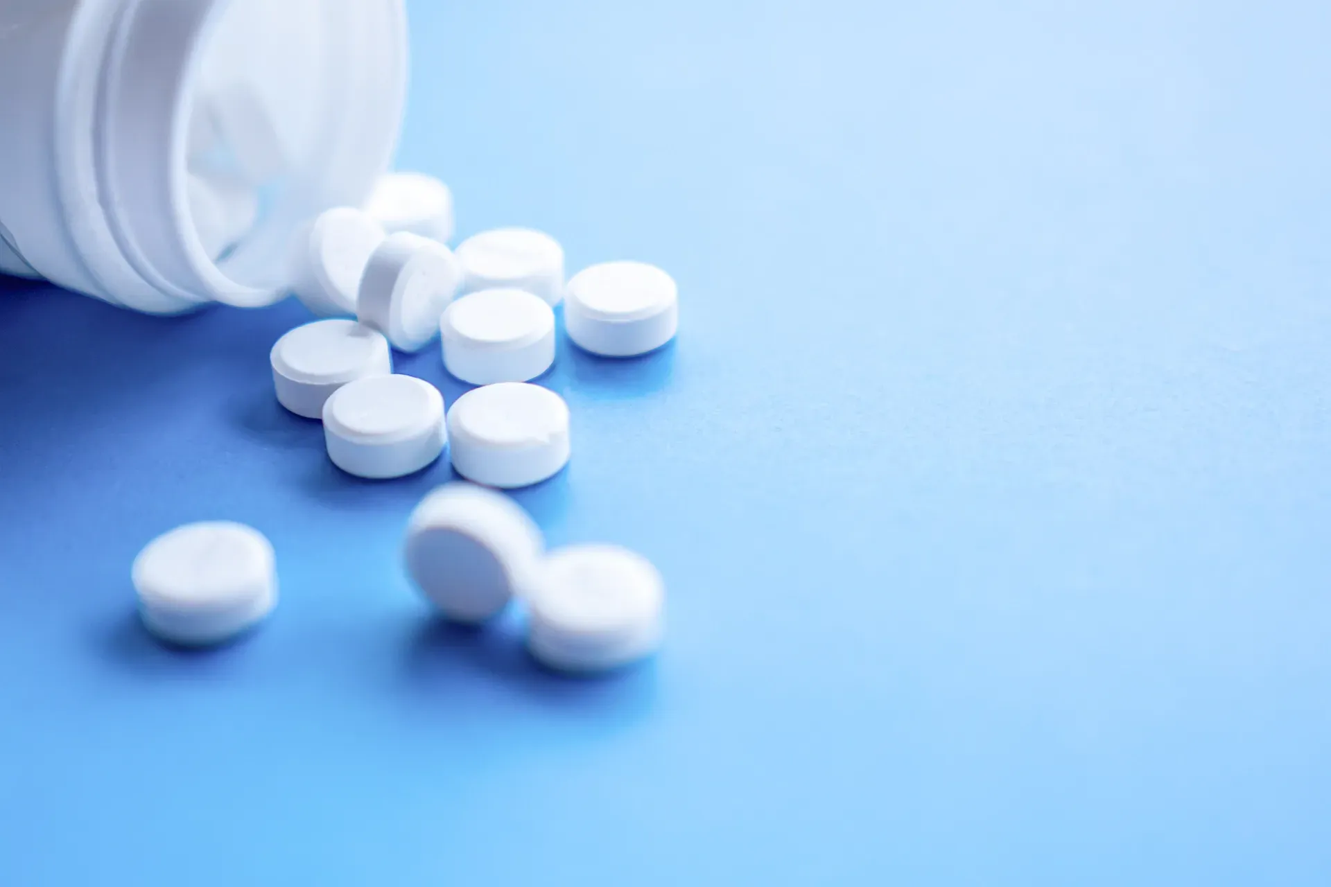 White pills are pouring out of a white bottle on a blue surface.