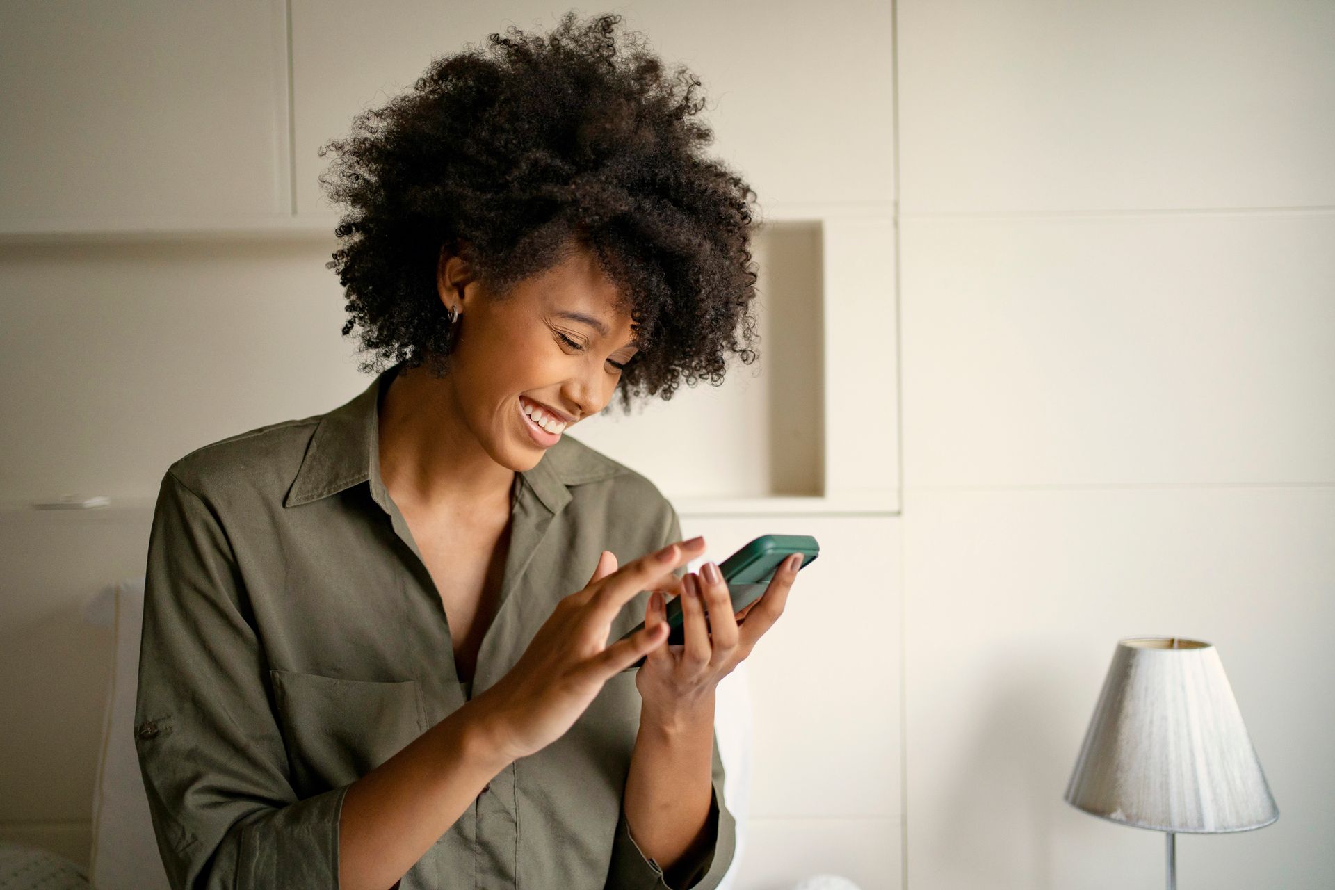 Woman with dark curly hair smiles while using a smartphone in a beige room.