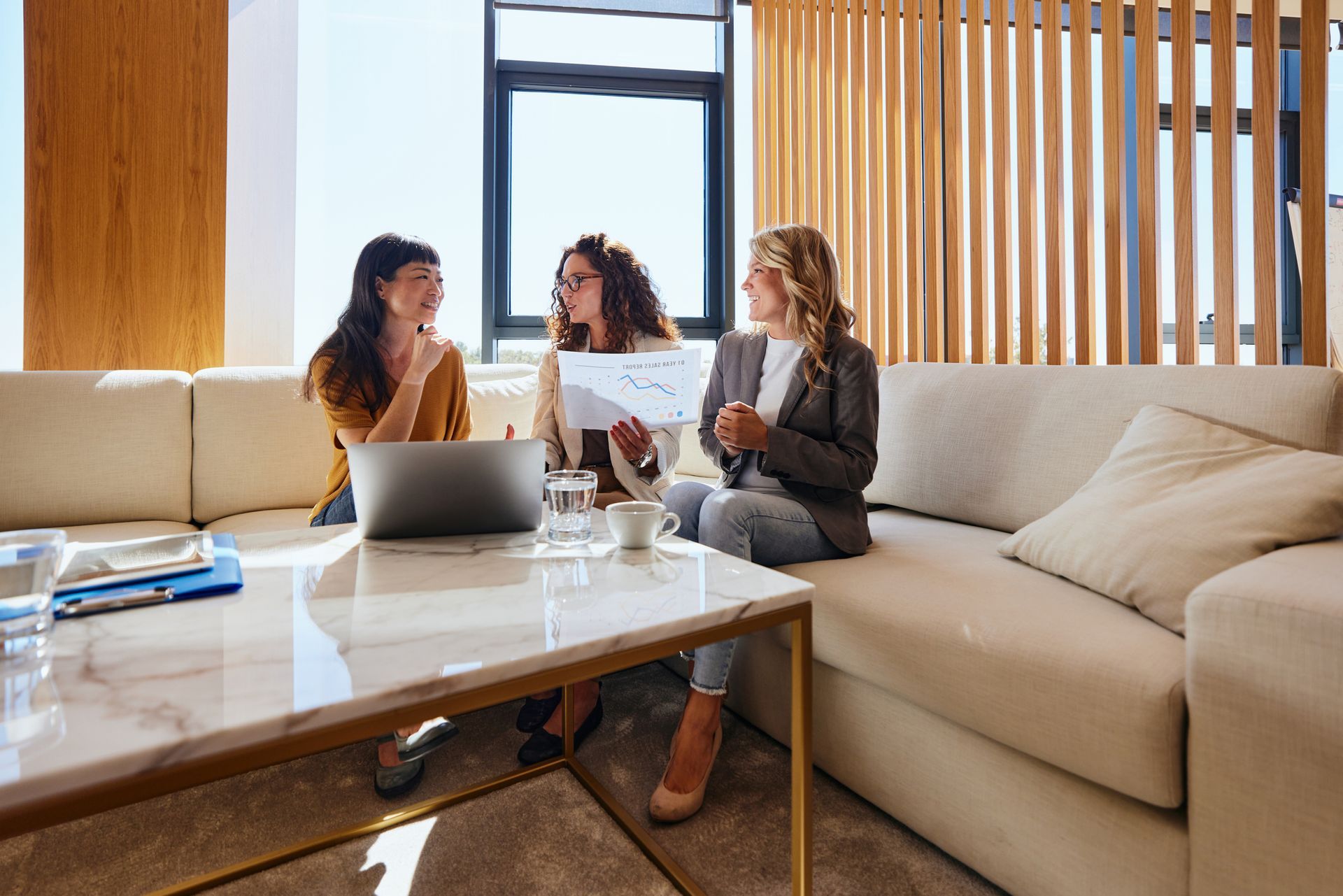 Three women in a modern office setting, reviewing documents, smiling.