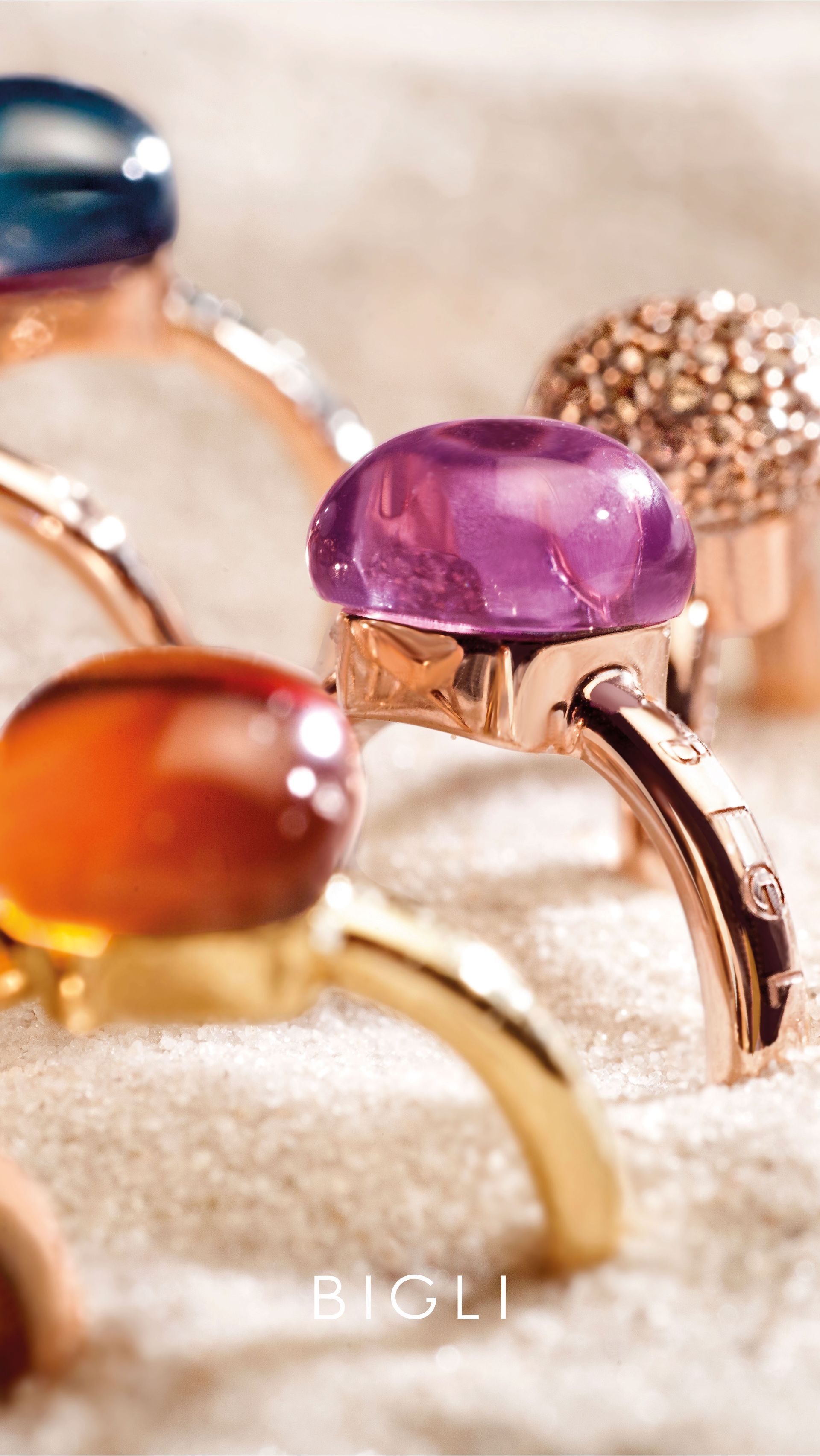 Nahaufnahme mehrerer Ringe mit bunten Edelsteinen, die auf Goldbändern gefasst sind und auf einer sandigen Oberfläche liegen.