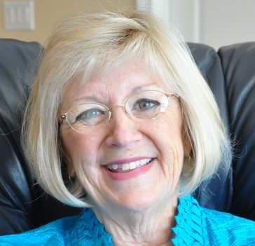 A woman wearing glasses and a blue shirt is smiling while sitting in a chair.