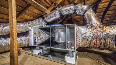 Large silver HVAC ductwork and equipment in an attic with exposed wooden beams and insulation.