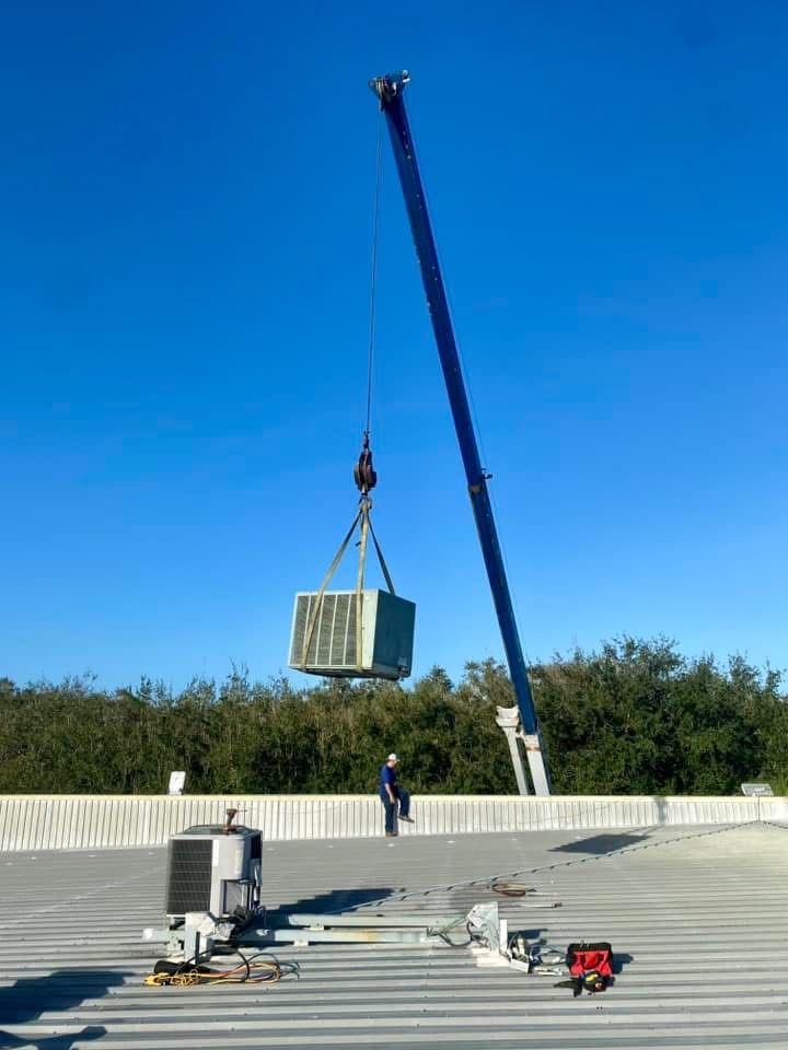 Crane lifting HVAC unit onto a rooftop. Person standing nearby, clear blue sky.