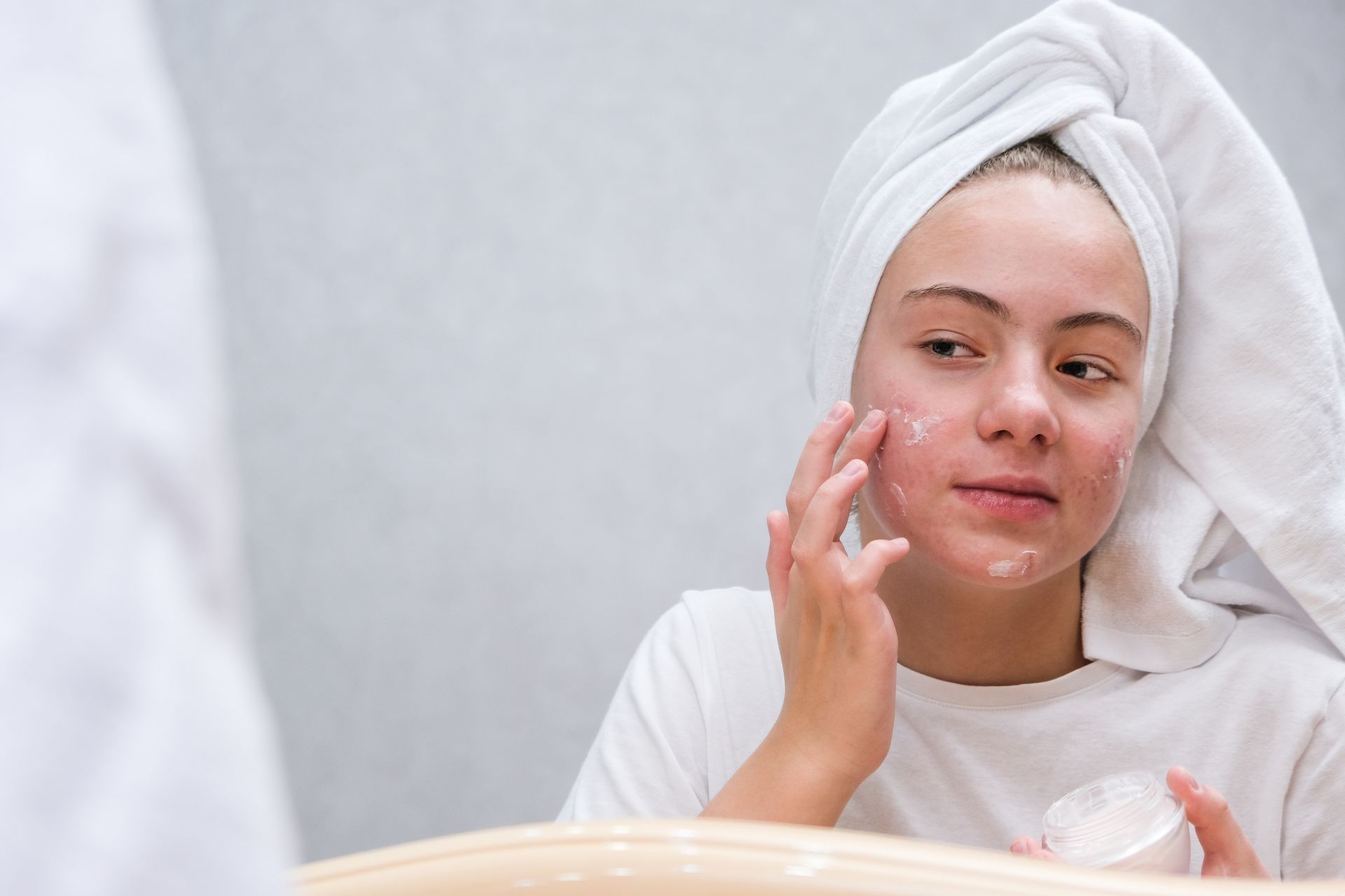 A woman with a towel wrapped around her head is applying cream to her face in front of a mirror.