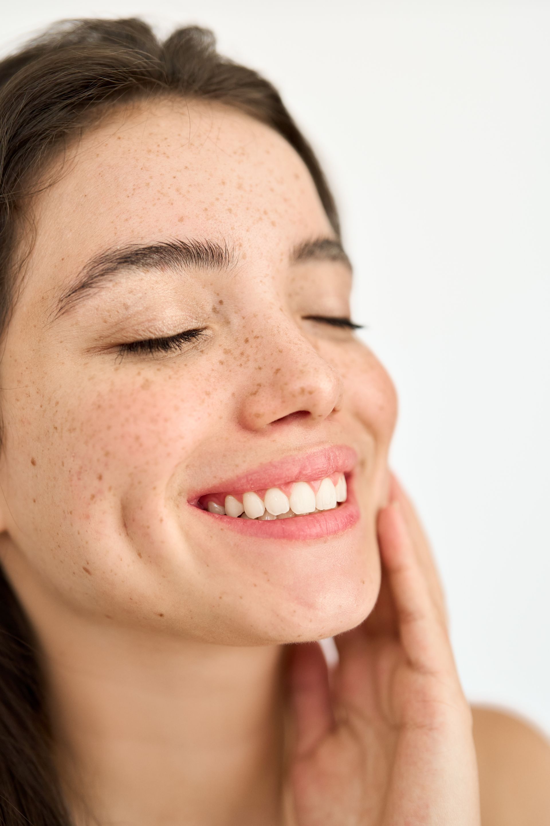 A woman with freckles is smiling with her eyes closed and touching her face.