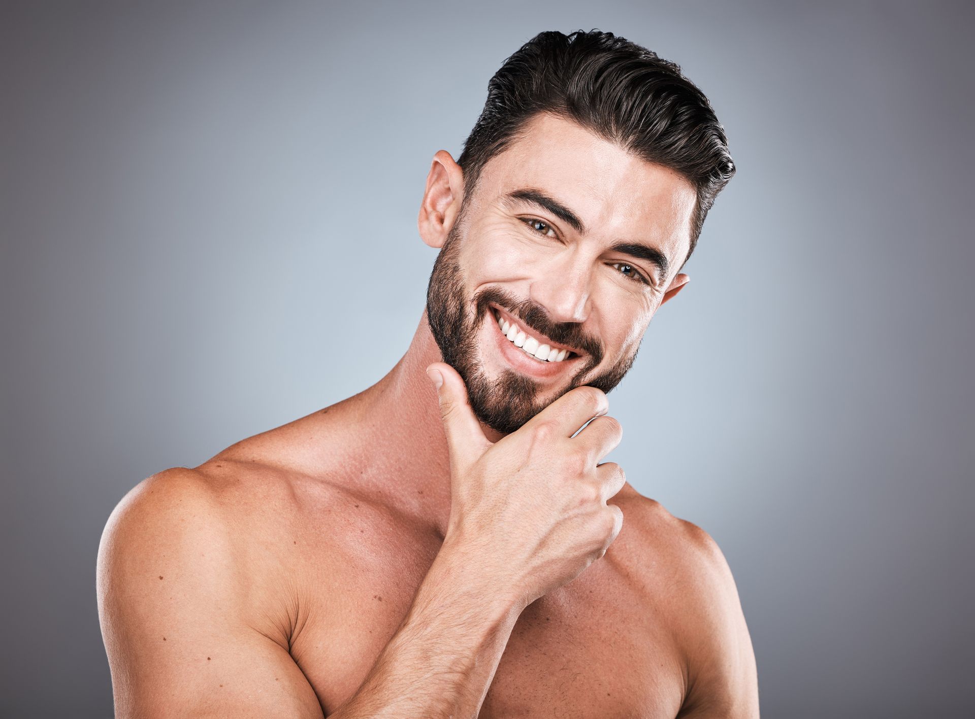 A shirtless man with a beard is smiling with his hand on his chin.