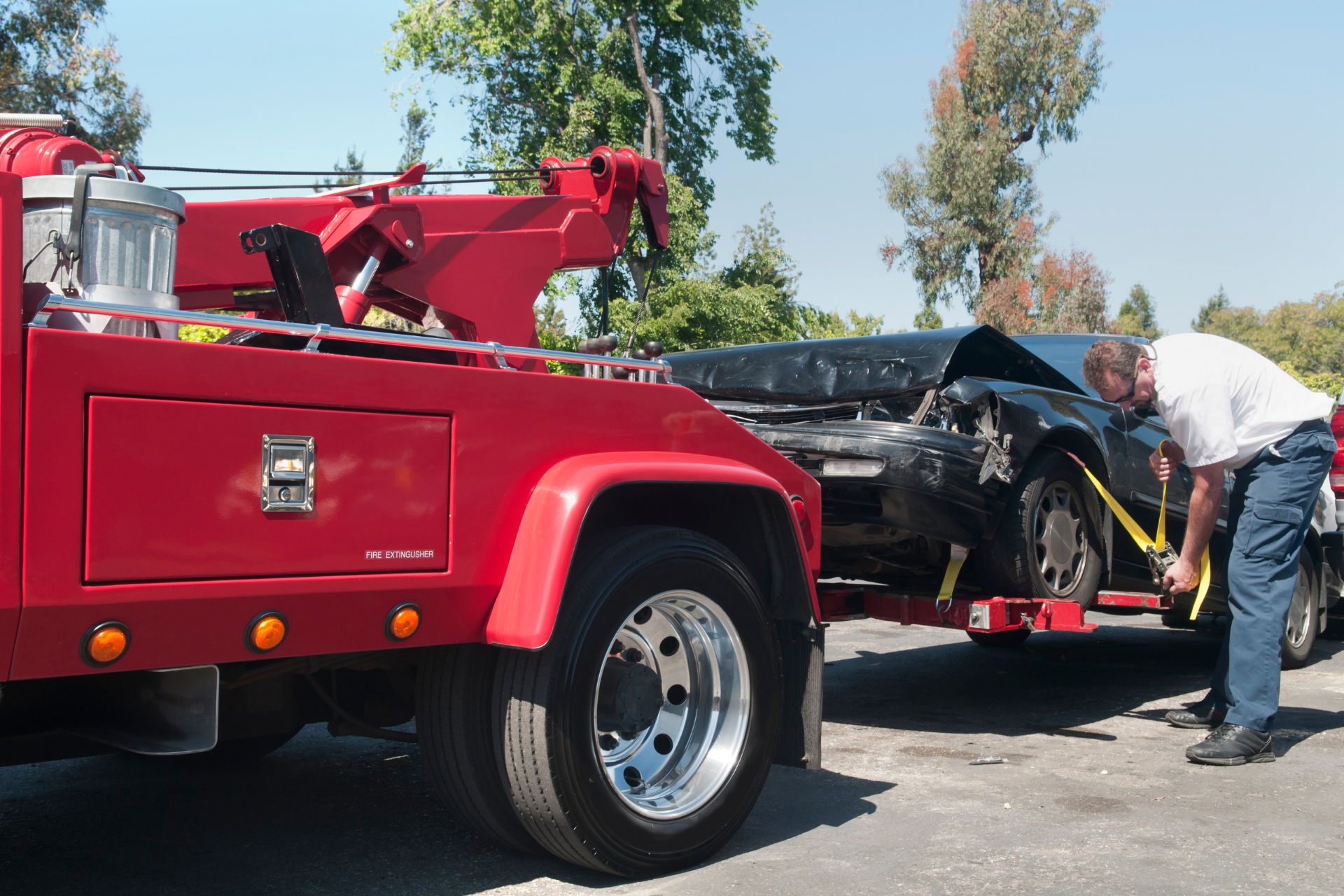 Red tow truck loading a black car while a man stands beside it on a sunny street