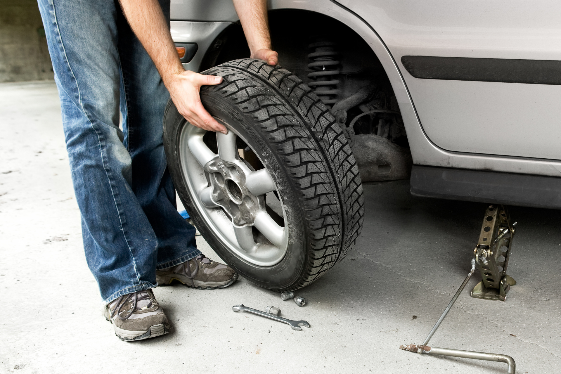 Person removing a car tire beside a silver vehicle with a jack on a driveway