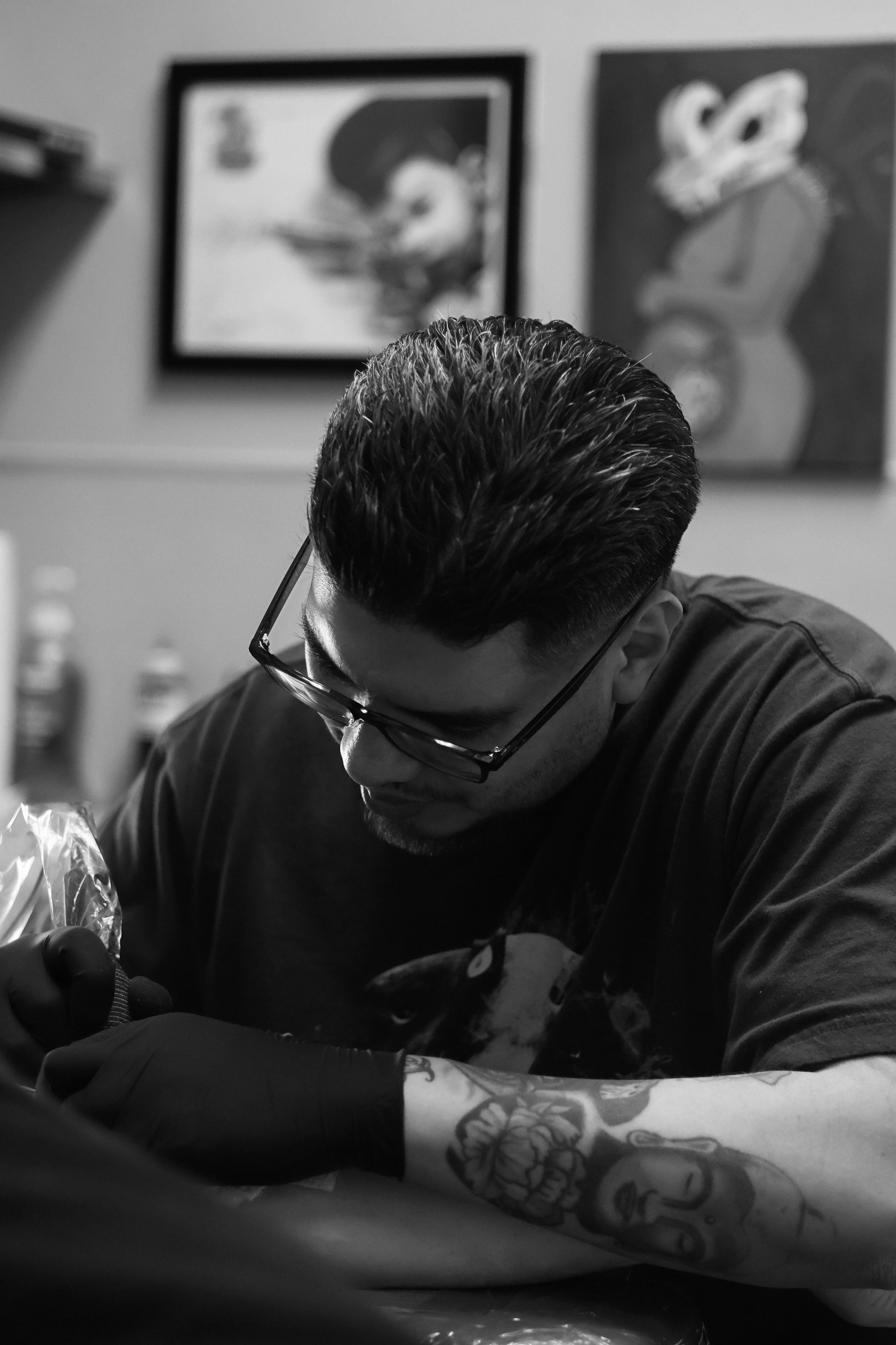 Tattoo artist working on an arm in a shop, wearing glasses and a dark shirt, in black and white.