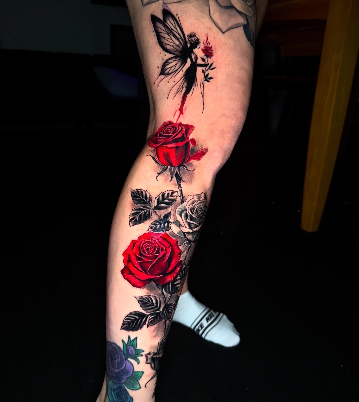Tattoo of a fairy with red roses on a leg, with black and gray leaves and black background.