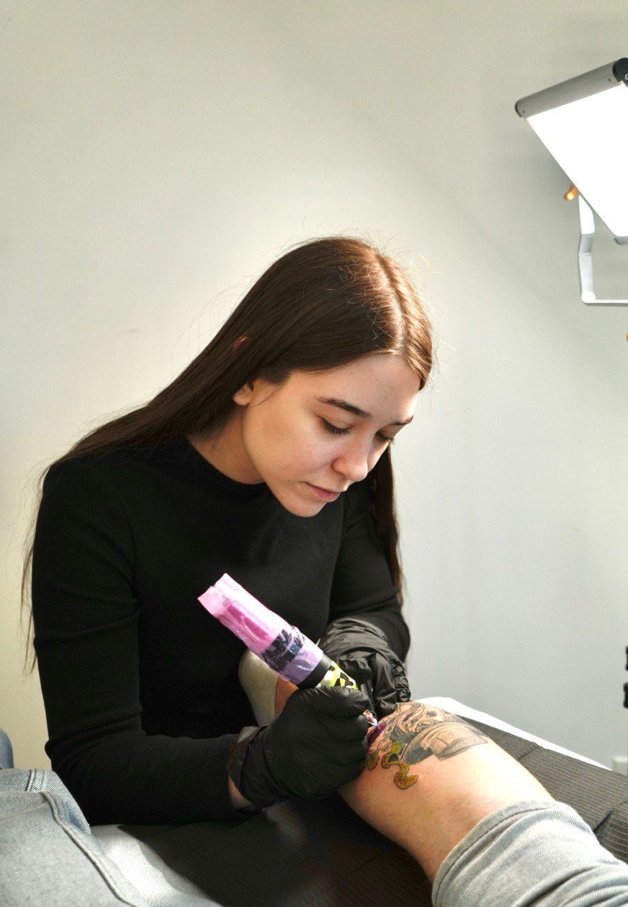 Woman in black shirt tattoos an arm, lit by a studio light.