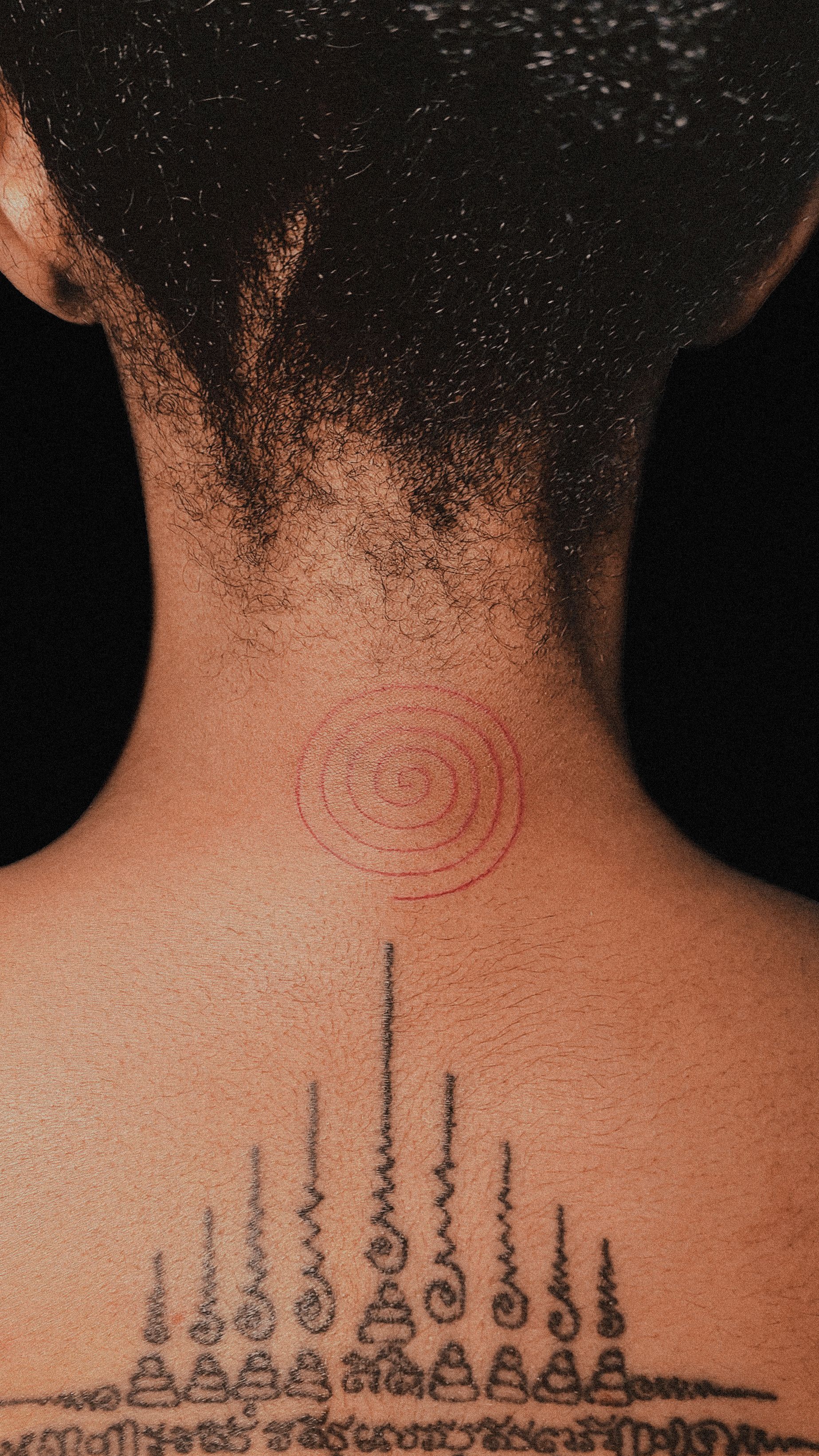 Back of a person with two tattoos: a spiral on the neck and a tiered design with script down the spine.
