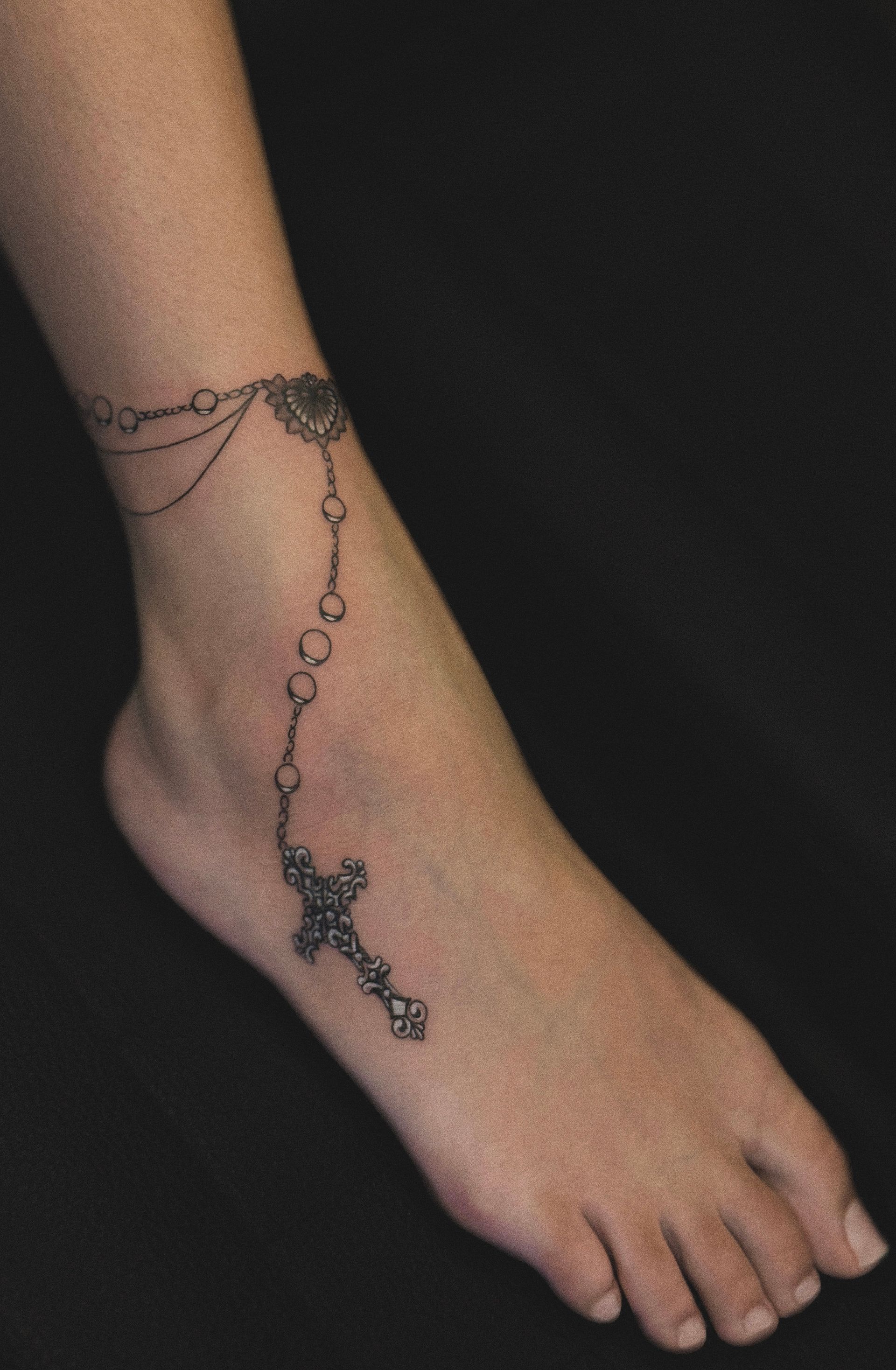 Ankle tattoo of a rosary with a cross pendant on a bare foot.
