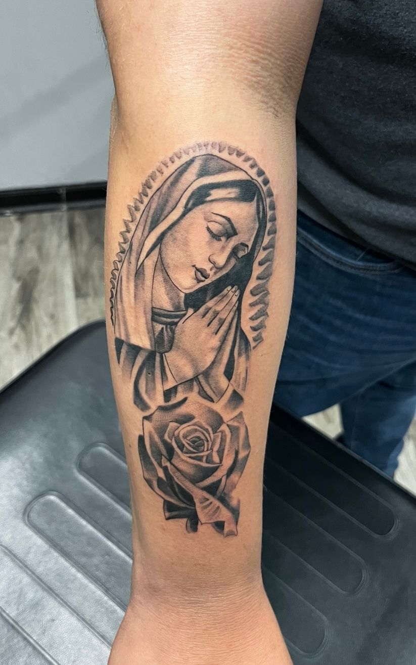 Black and gray tattoo of Virgin Mary praying, with a rose on arm.