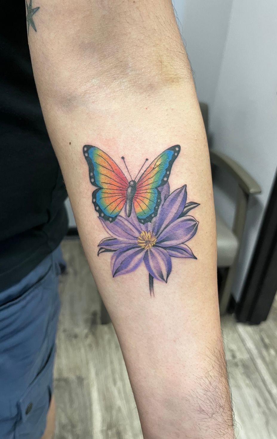 Tattoo of a colorful butterfly atop a purple flower on a person's forearm.