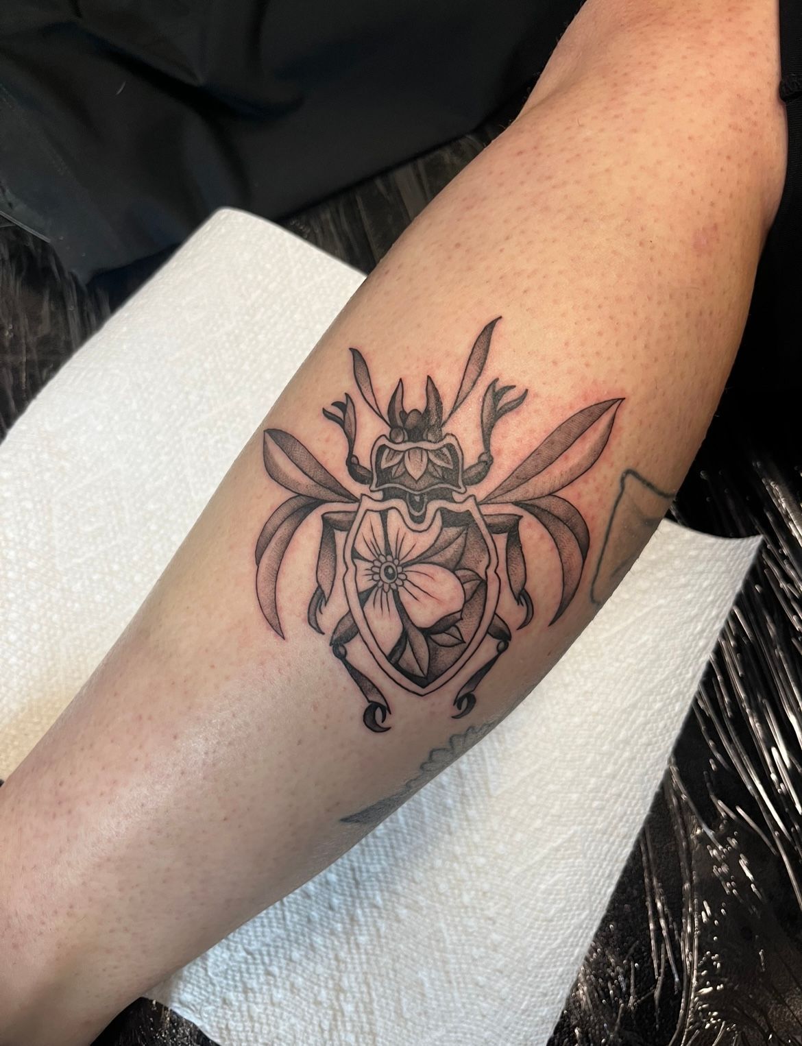 Black beetle tattoo on a person's arm; wings spread, flower within body.
