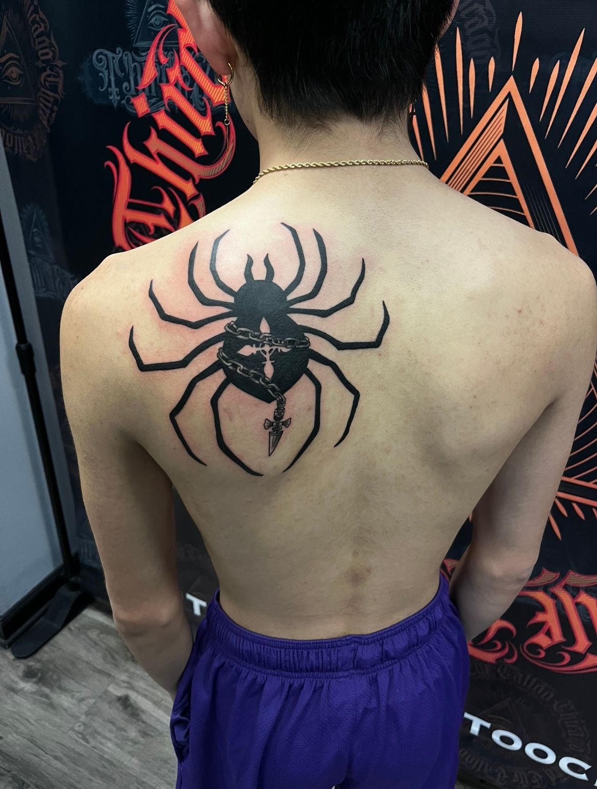 Person with back tattoo of a black spider with a skull-like design. They are wearing purple shorts.