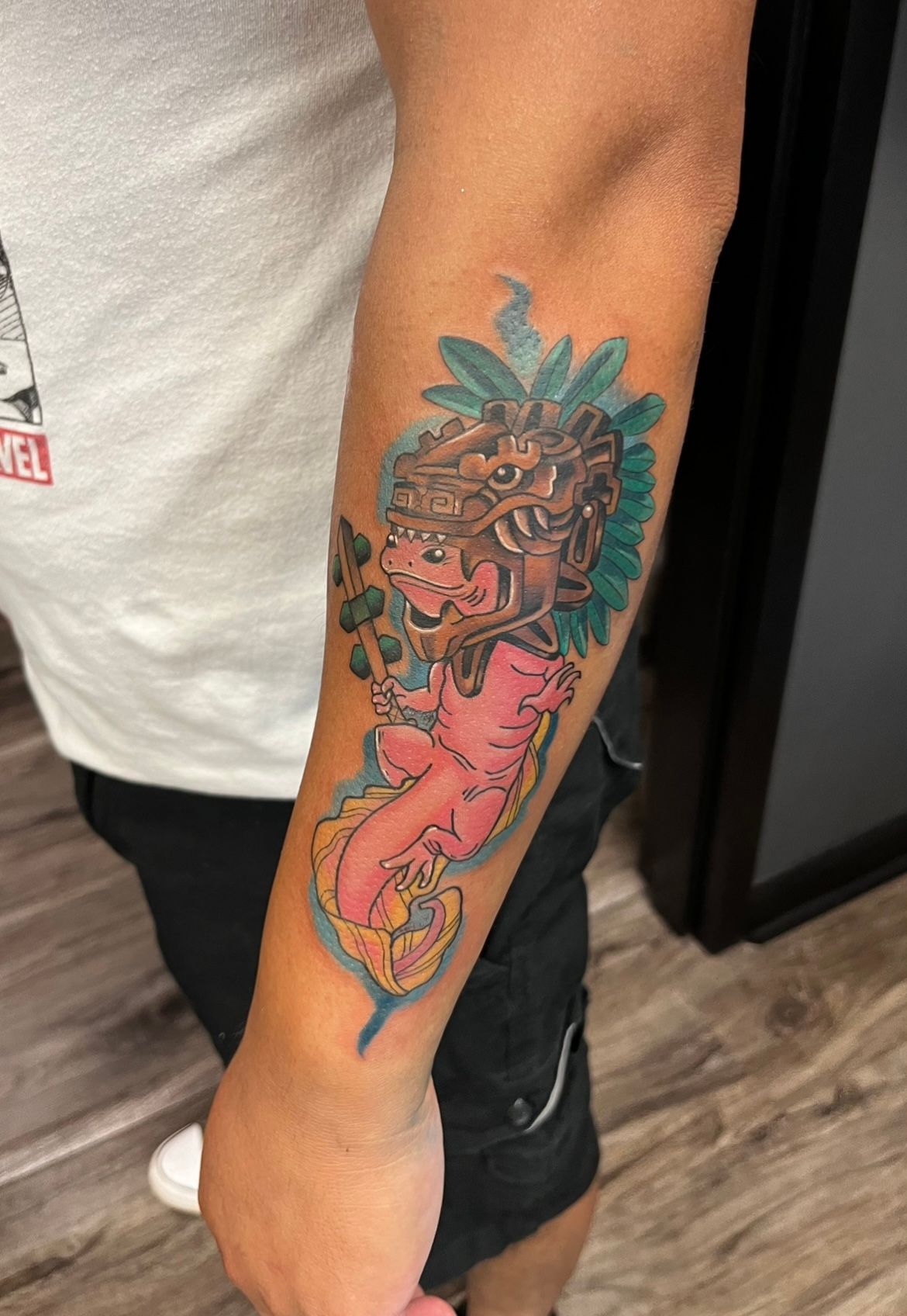 Arm tattoo of a pink Aztec figure with a detailed headdress, surrounded by blue and green foliage.
