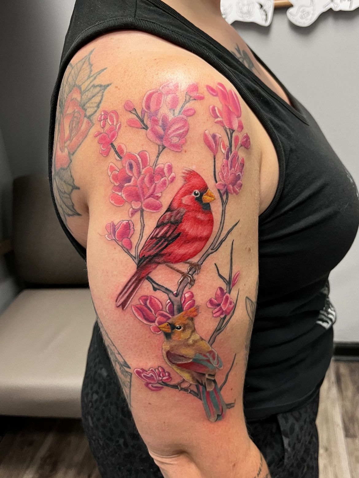 Tattoo of two cardinals perched on a flowering branch. Red and pink colors. On a person's upper arm.