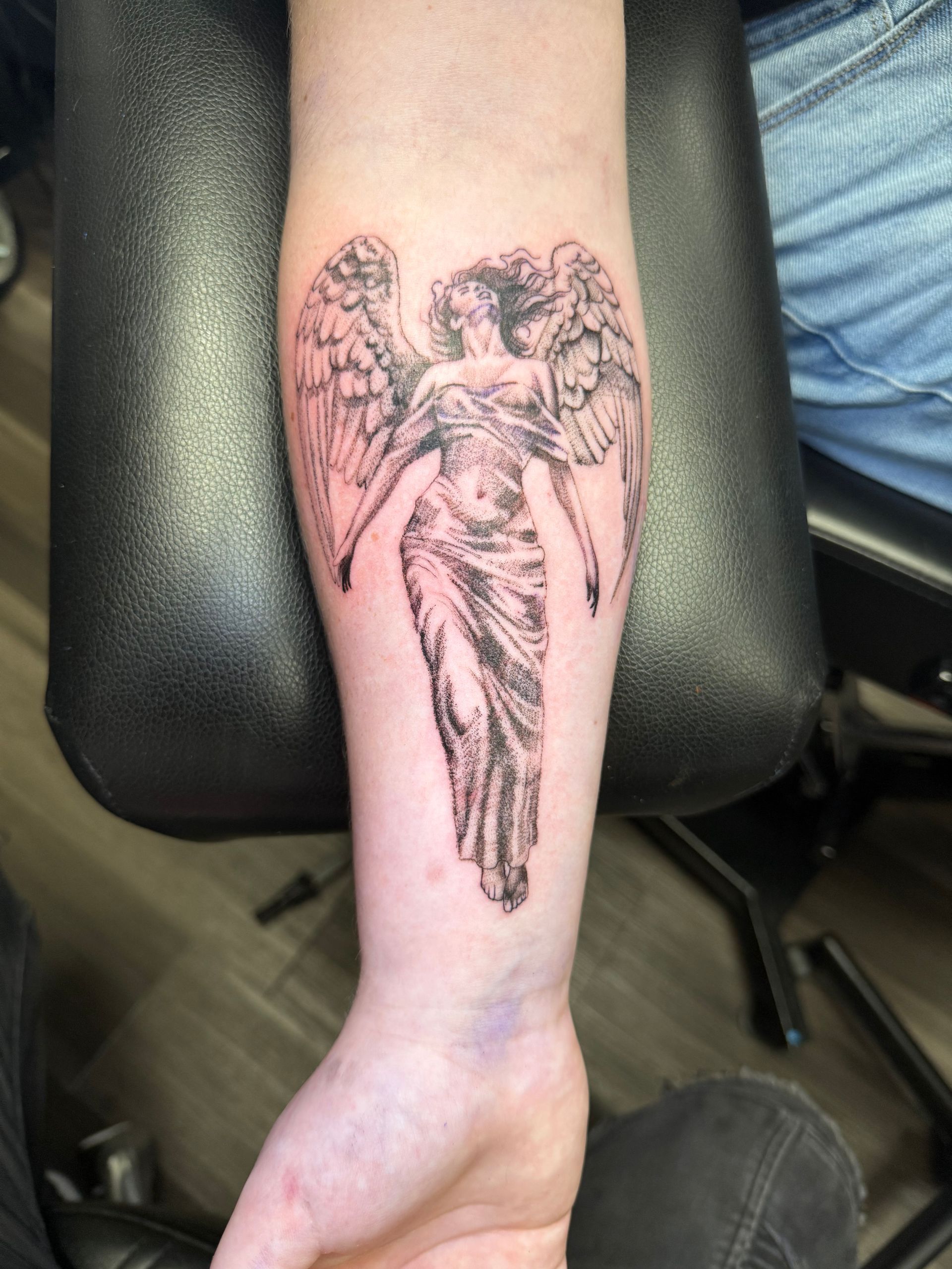 Tattoo of a winged angel on a person's forearm. Gray shading and detailed sculpture style.