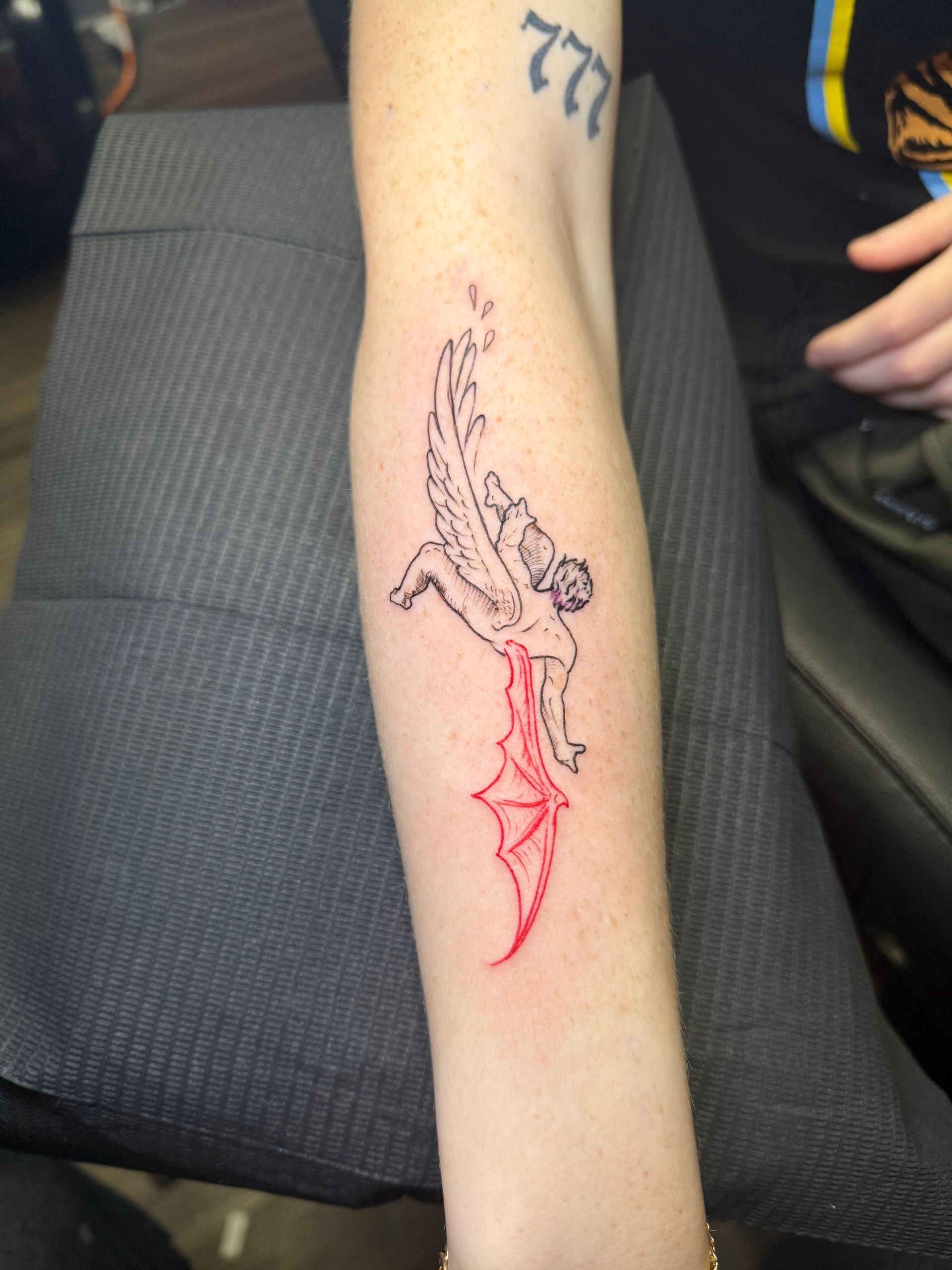 Tattoo of an angel falling, black silhouette with red accents, on forearm, arm resting on grey surface.