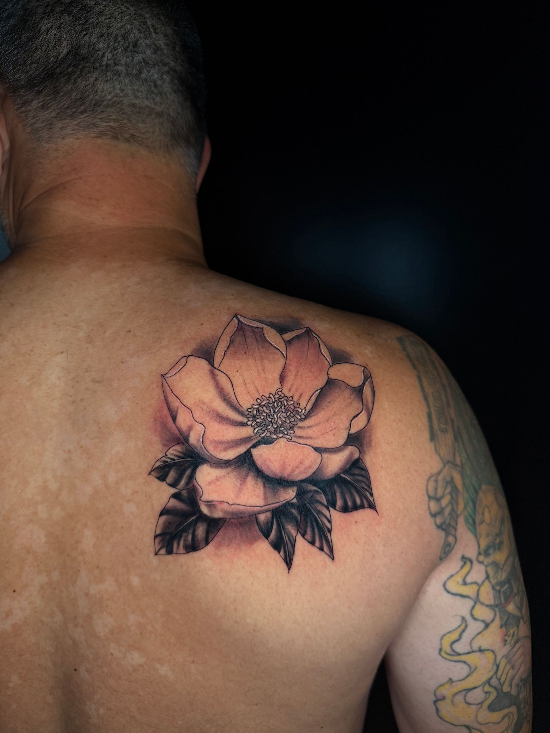 Tattoo of a peach-colored flower with dark leaves on a man's back and shoulder.