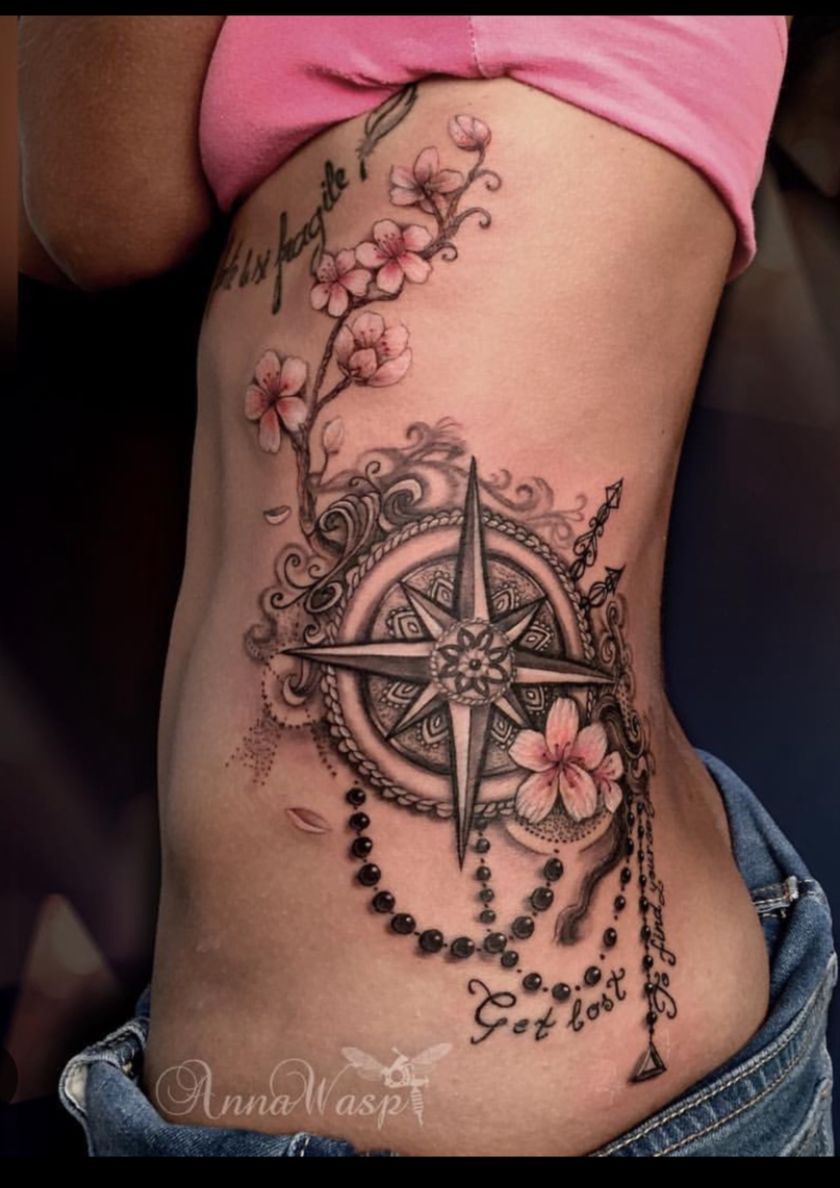 Tattoo on side featuring a compass rose, floral branch, and beaded strings. Black and gray ink.