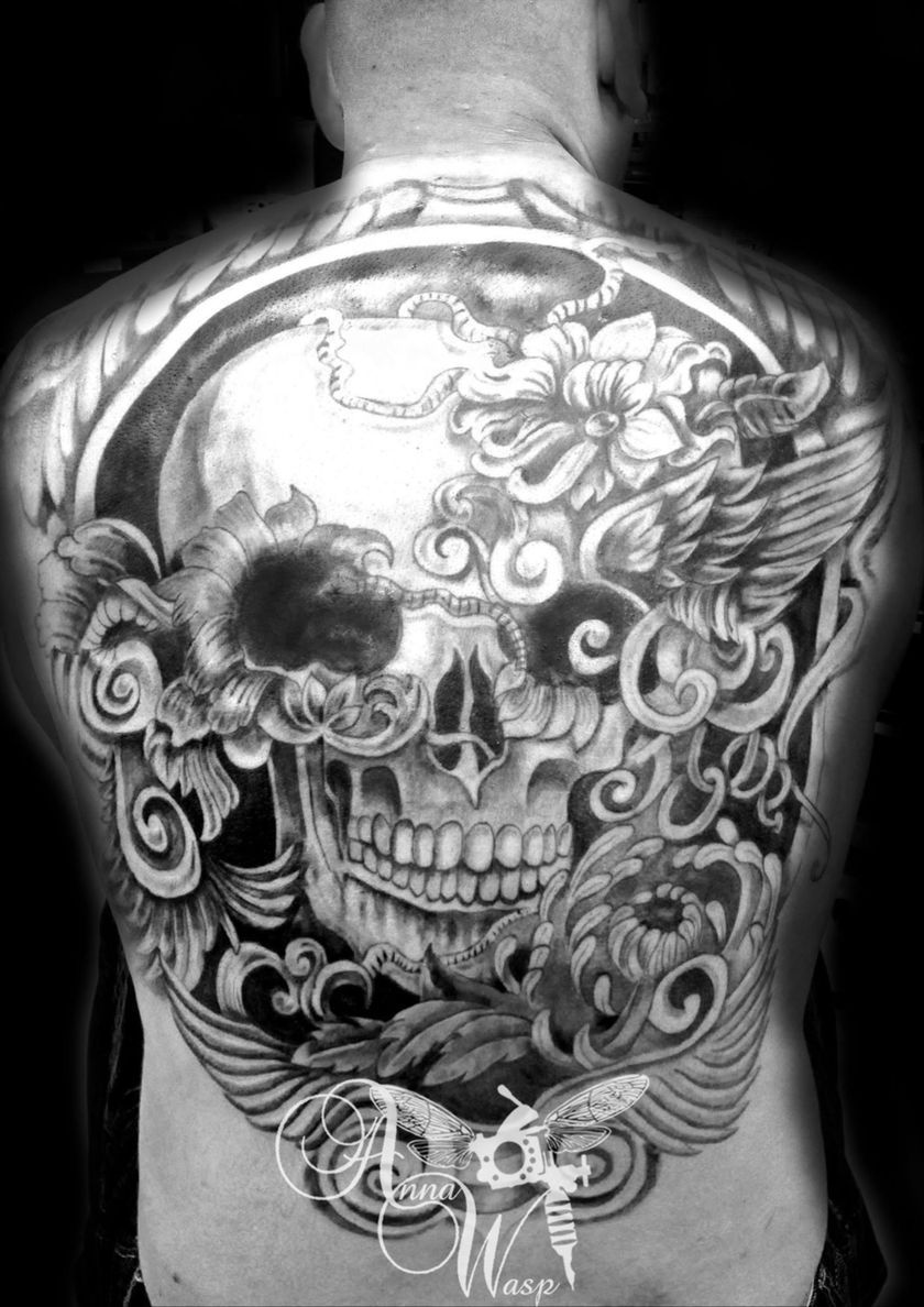 Black and white back tattoo of a skull with floral and wing designs inside a circular frame.