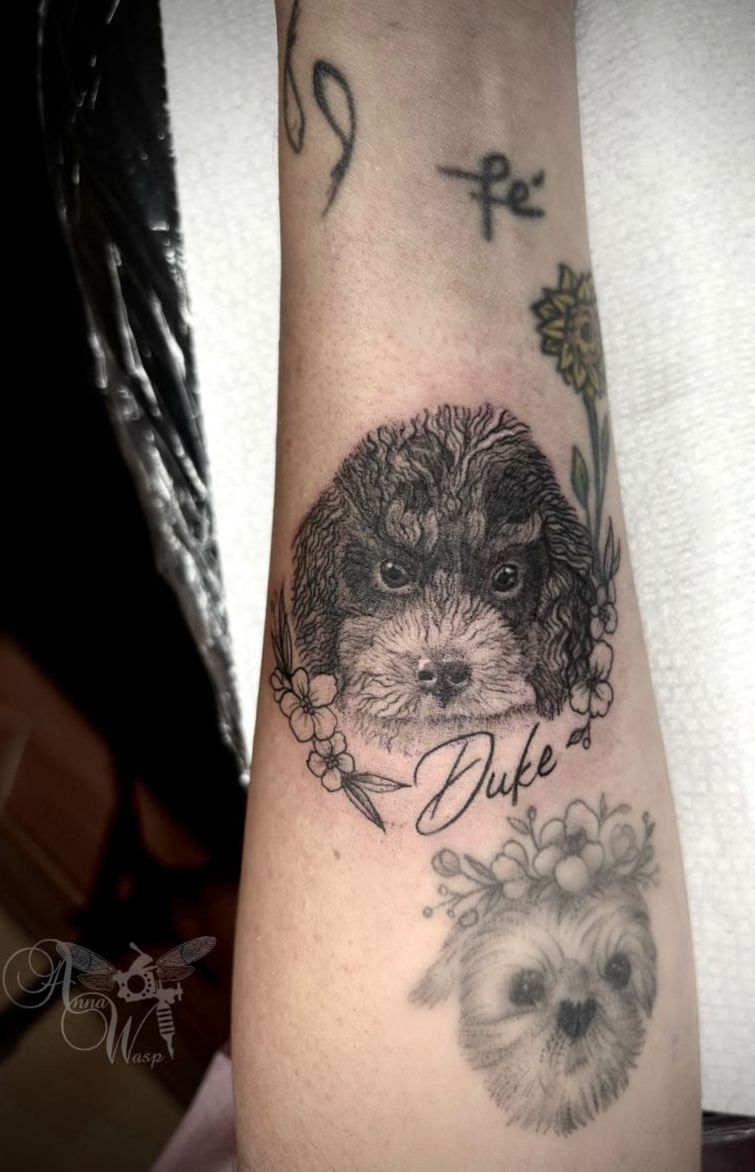 Tattoo of a dog named Duke with floral accents, and another faded dog tattoo below on an arm.