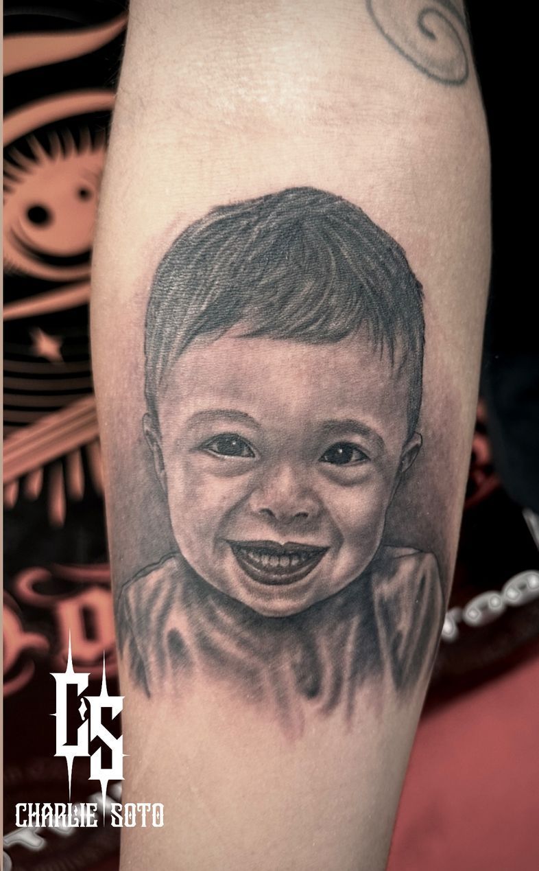 Black and white portrait tattoo of a smiling toddler on a person's forearm.