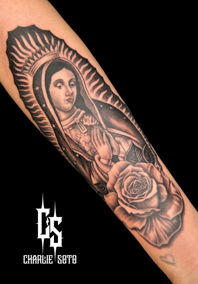 Black and gray tattoo of Virgin Mary with halo and rose on arm.