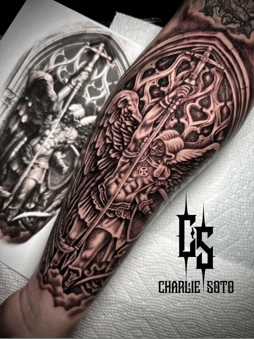 Tattoo of Archangel Michael with sword and shield inside Gothic window. Black and grey, detailed, on an arm.