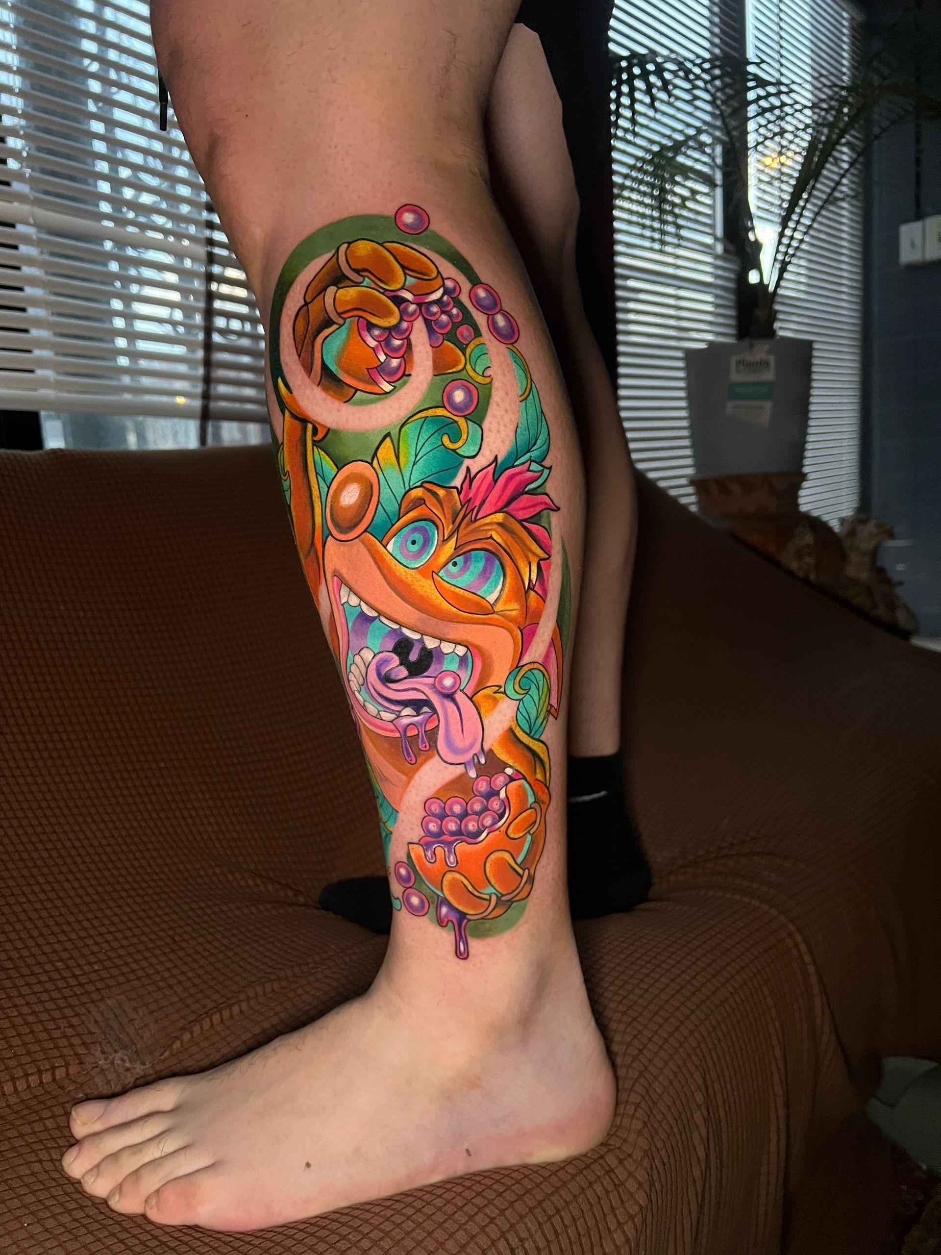 Tattoo on calf: cartoon character with tongue out, surrounded by colorful swirls and foliage, on a leg.