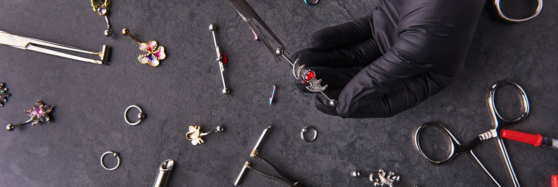 A black-gloved hand holding jewelry with surgical tools scattered on a dark surface.
