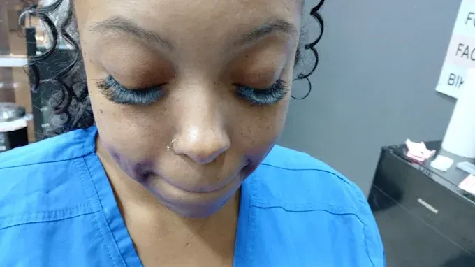 A person in blue scrubs with dark eyelashes and purple markings on face, looking down.
