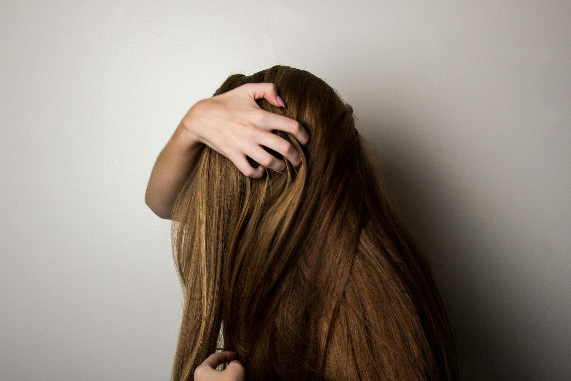 Woman with long brown hair touching her head against a plain white wall.