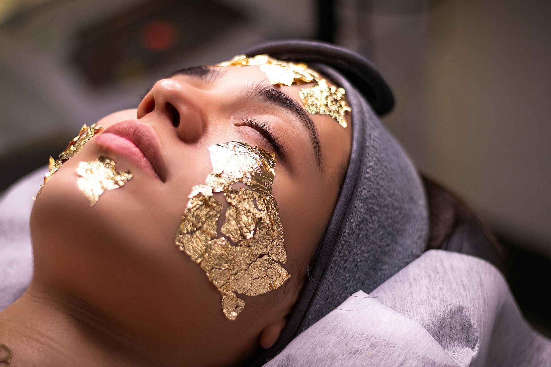 Woman with gold leaf facial treatment, eyes closed, resting on a spa bed, light skin tone.