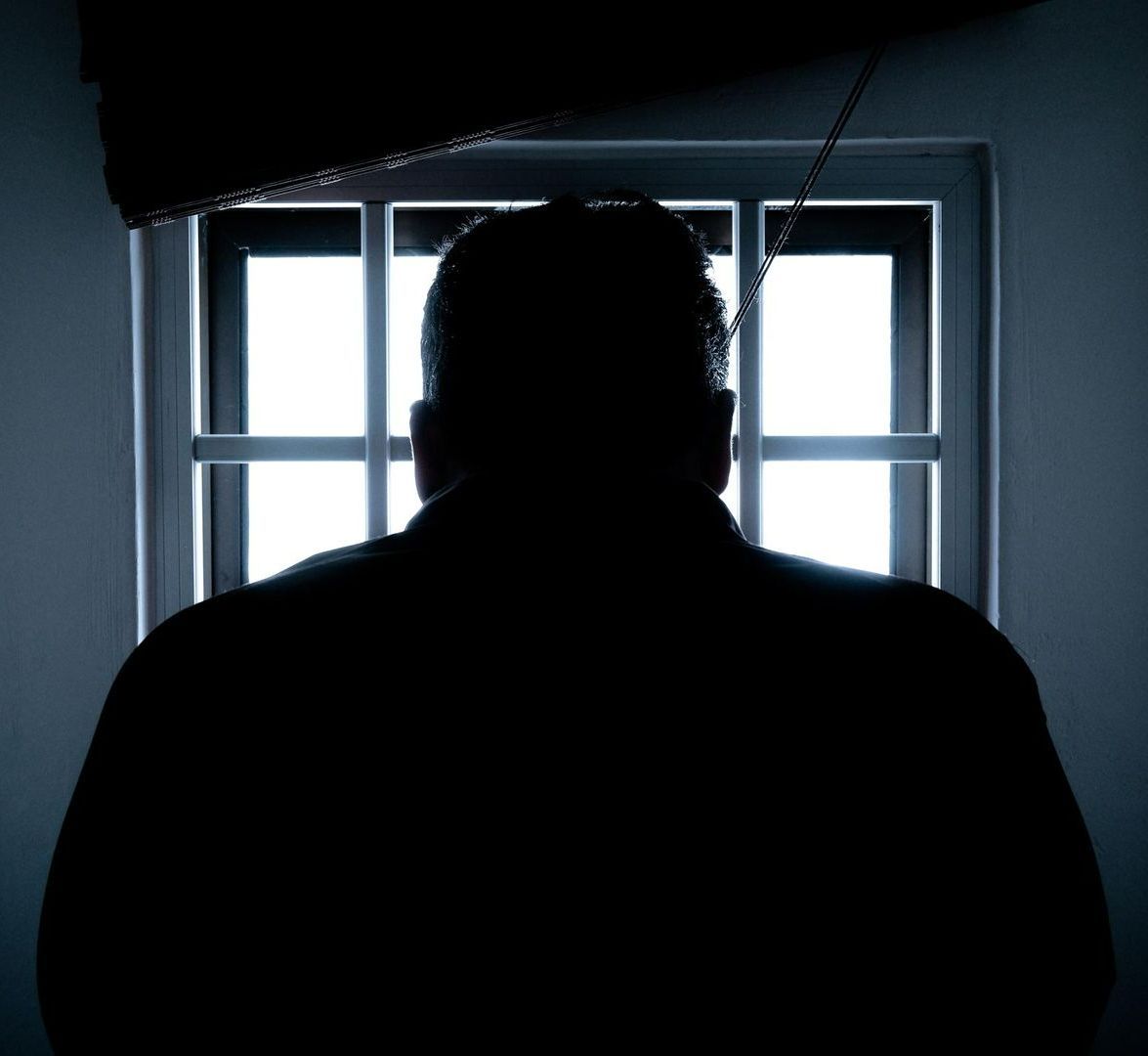 A silhouette of a man behind bars looking out of a window.