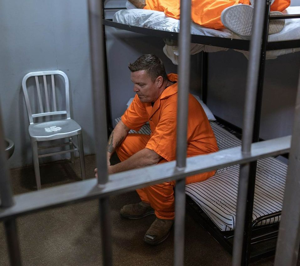 A man in an orange jumpsuit is sitting on a bunk bed in a prison cell.