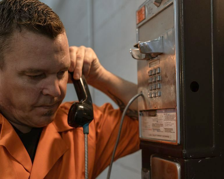 A man in an orange shirt is talking on a pay phone