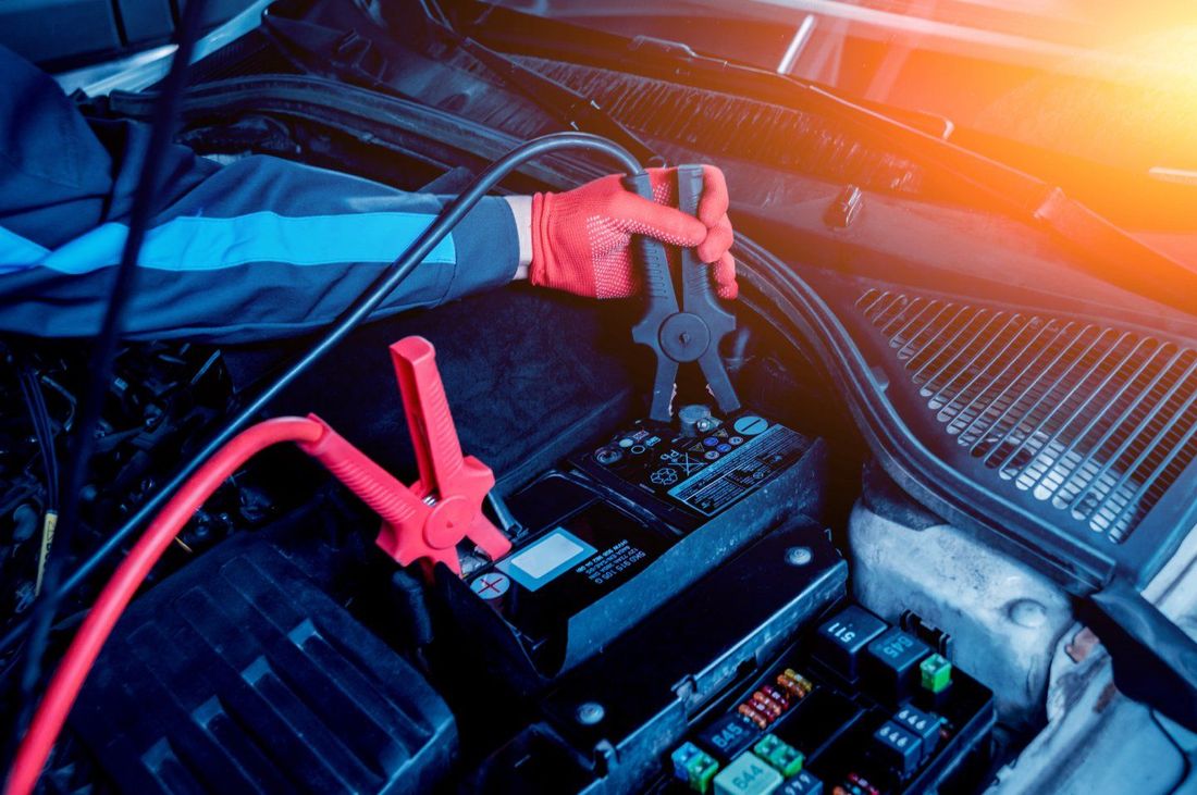 Una mano guantata fissa un morsetto nero per cavi di avviamento a un terminale della batteria di un'auto, con un morsetto rosso visibile nelle vicinanze.