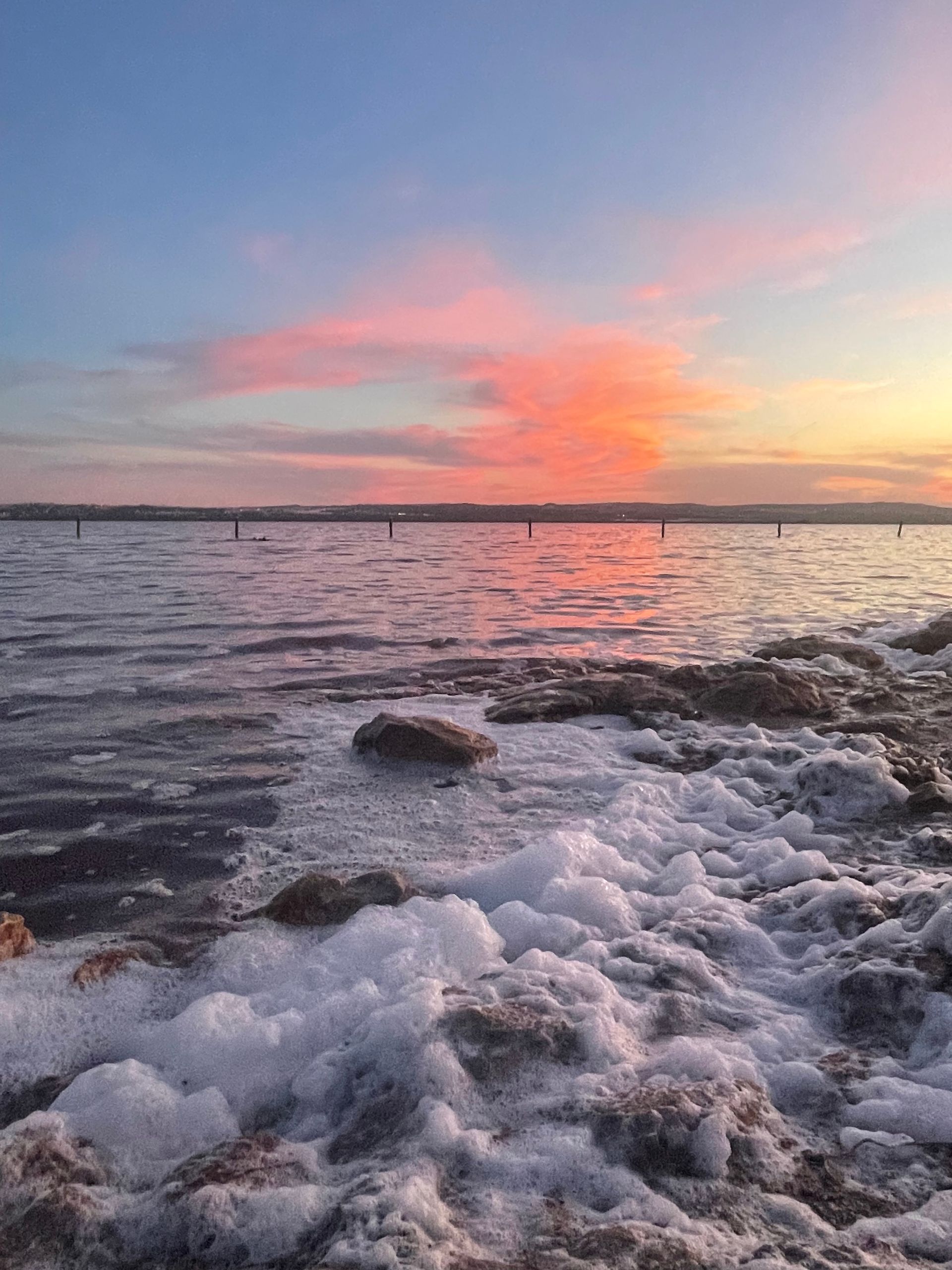 Een zonsondergang boven een watermassa met rotsen op de voorgrond en een brug op de achtergrond.