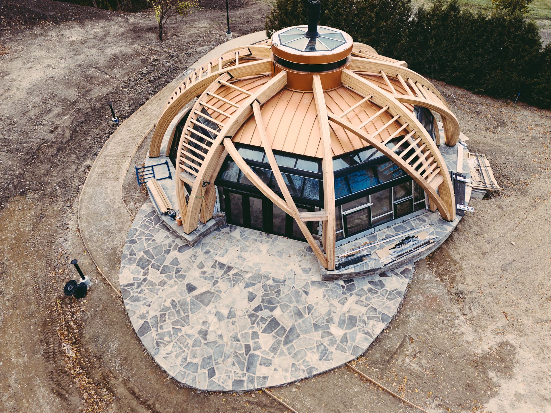 Aerial view of a wooden geodesic-style pavilion with a stone patio on a dirt site