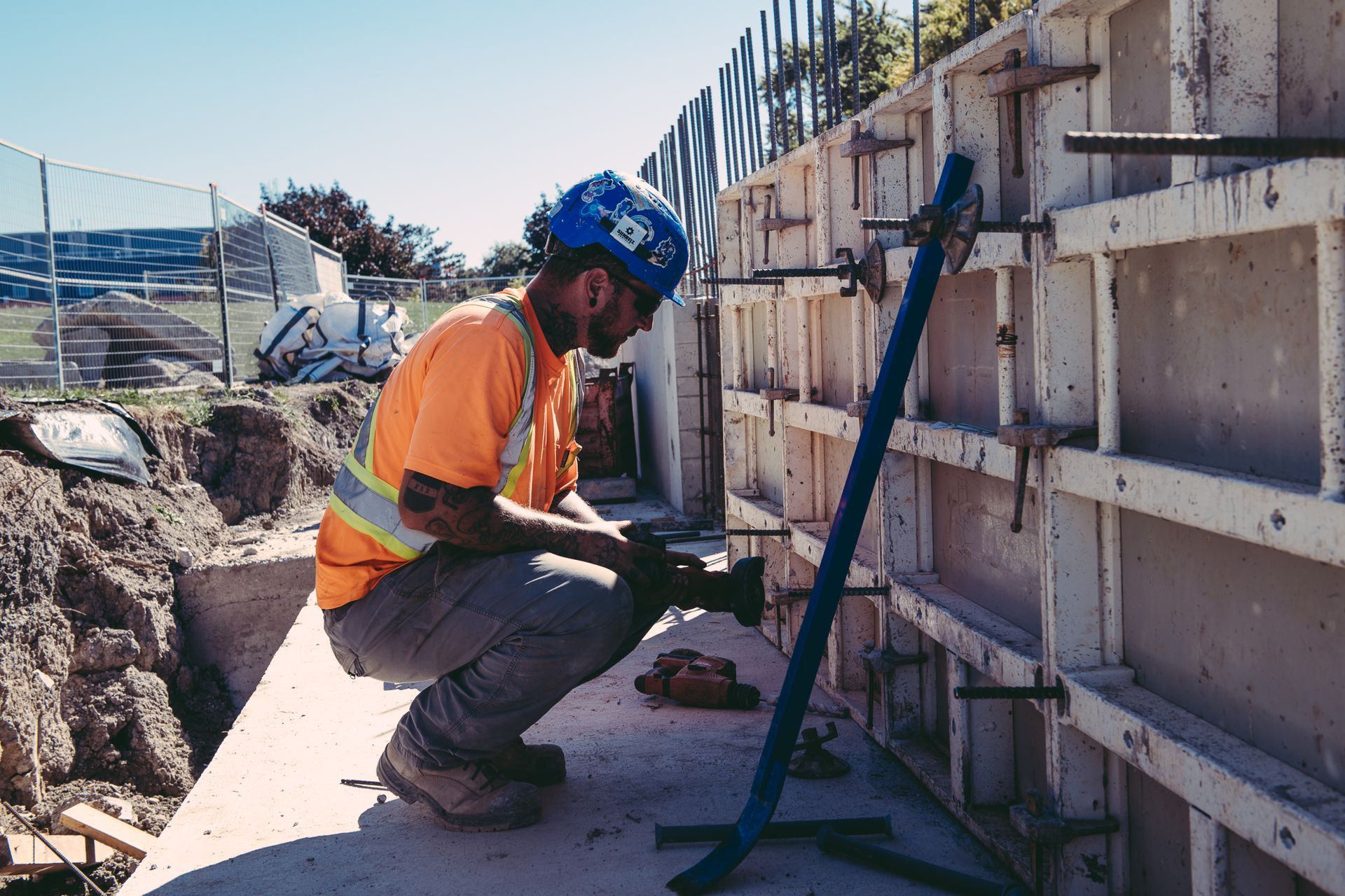 Construction worker kneeling beside a concrete wall, inspecting a blue strap at a worksite.