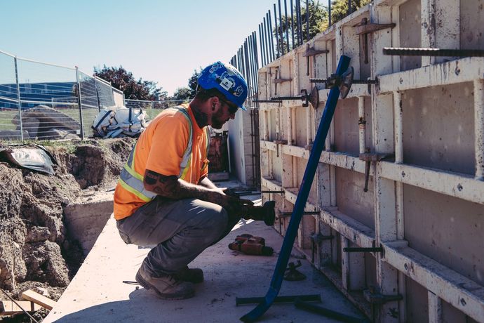 Construction worker in orange vest and blue hard hat crouches beside a concrete wall at a work site.