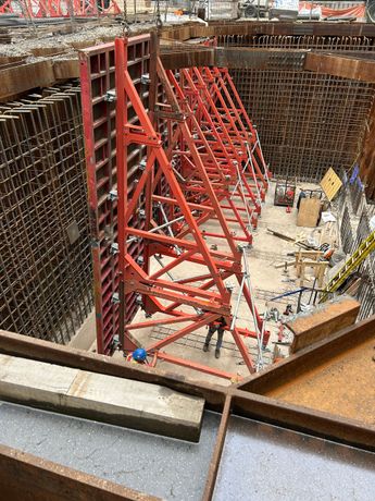 Red steel bracing inside a deep construction pit with workers and scaffolding along the walls