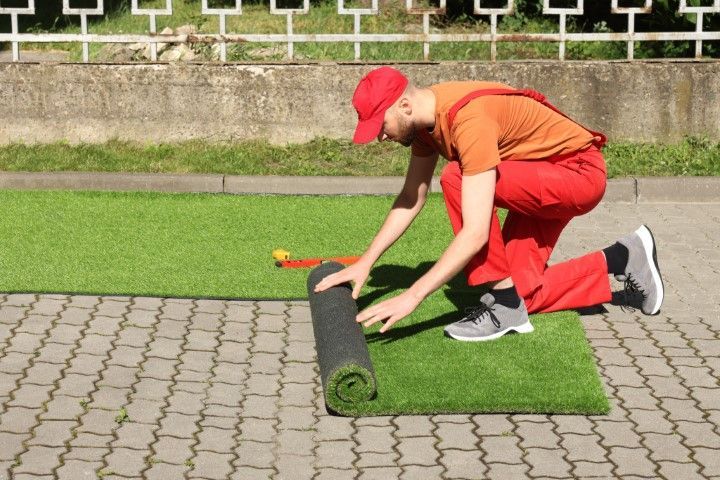 A person in orange clothes and a red cap kneeling on a paved surface to unroll a strip of artificial green grass.
