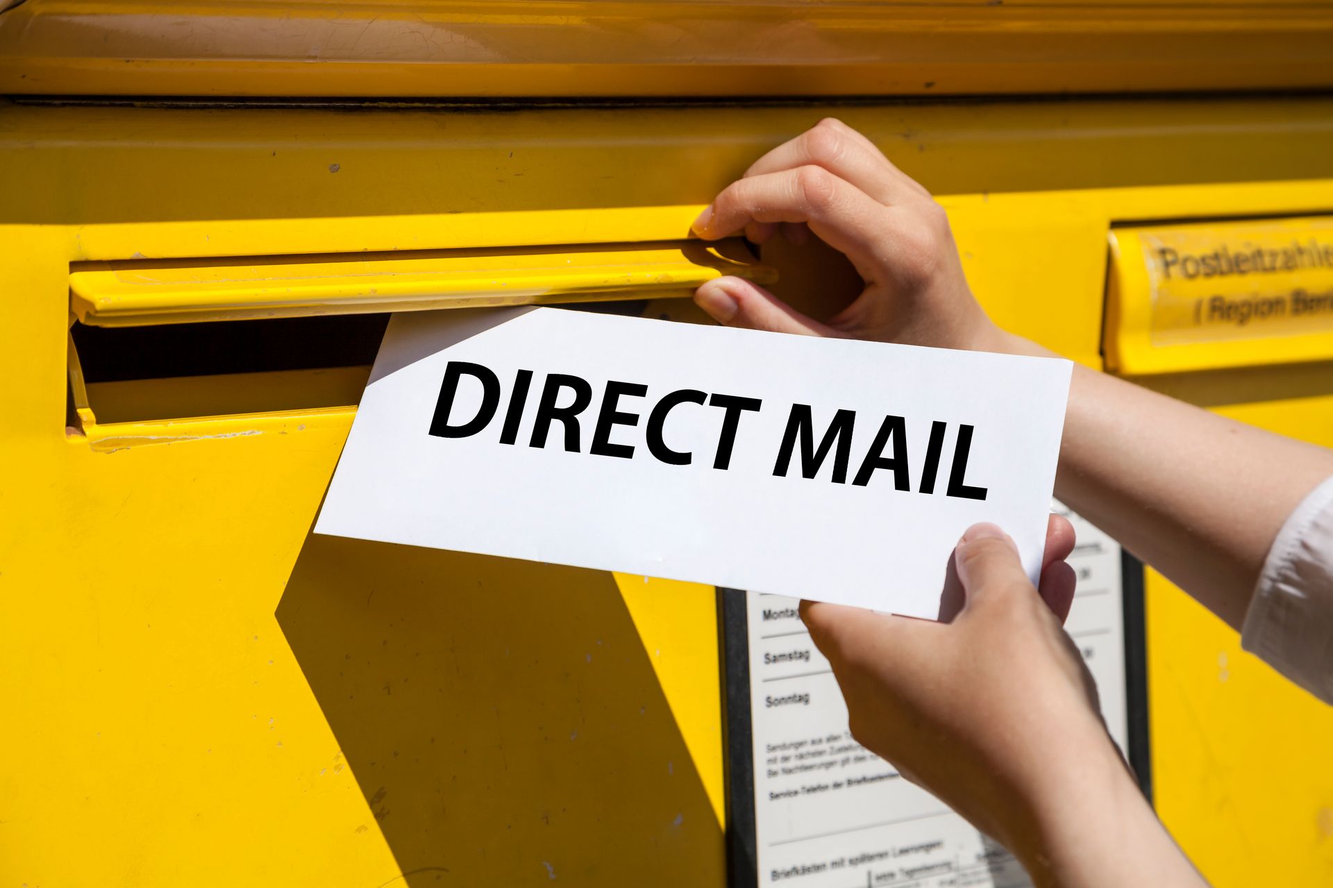 A person is putting a direct mail envelope into a yellow mailbox