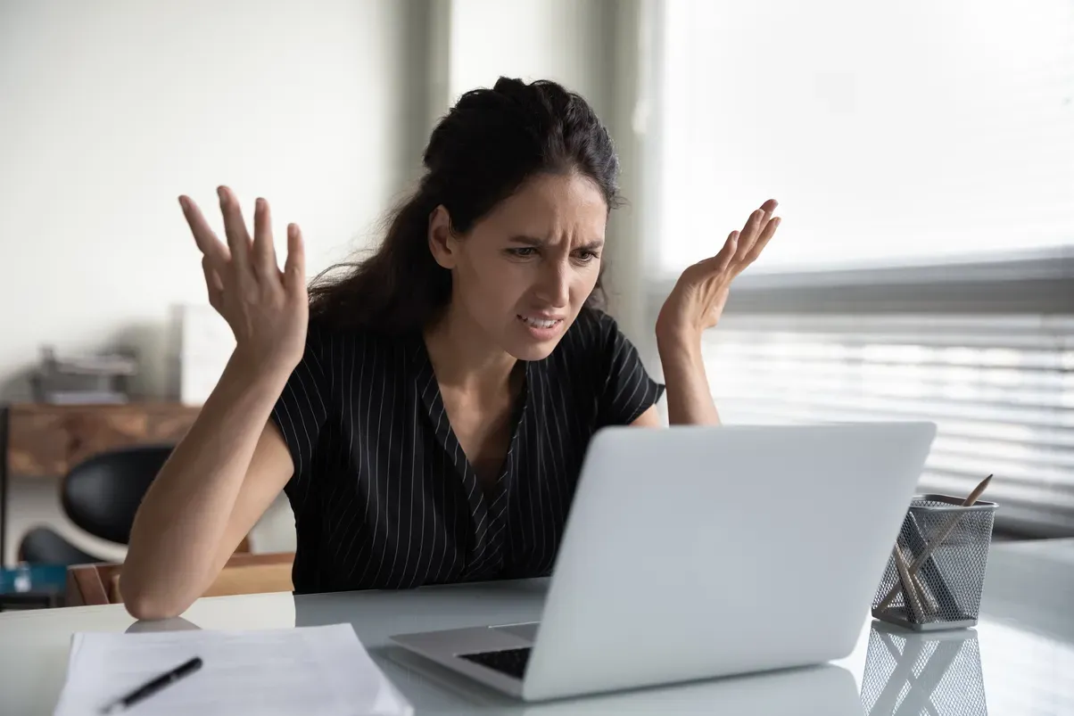Woman at a desk with a laptop, looking frustrated with arms raised, office setting.